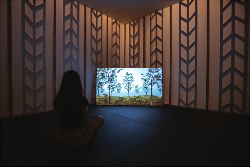 A person seated on a gallery floor with their back turned, facing a corner where a screen projects an image of a forest.